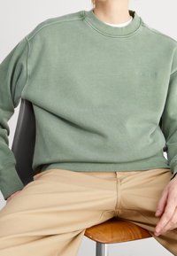 Felpa verde con colletto e polsini a coste, con un piccolo logo ricamato. Abbinata a pantaloni beige, seduto su una sedia di legno.