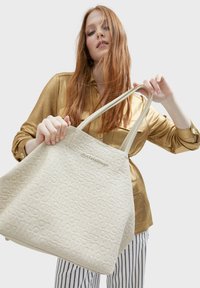 Beige toteväska med präglat logomönster, textilstruktur och långa handtag. Modellen håller den framför en neutral bakgrund.