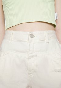 Gros plan d'une personne portant un crop top côtelé vert clair et un pantalon taille haute écru avec fermeture par boutons.