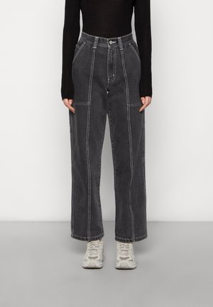 Person trägt ein schwarzes Langarmshirt, hoch taillierte weite dunkelgraue Jeans mit weißen Nähten und weiße Sportschuhe und steht auf grauem Boden.
