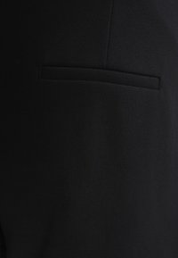 Tissu noir à la texture lisse avec une petite poche verticale horizontale. Le design global est minimaliste et ajusté.