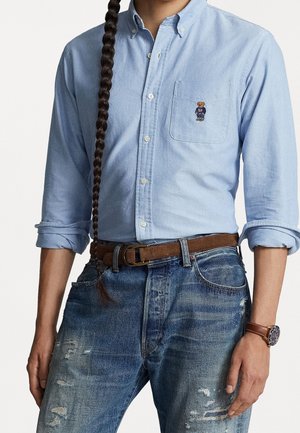 Homme portant une chemise bleu clair à boutons avec un logo d'ours sur la poche, un jean bleu usé, une ceinture marron et une montre-bracelet.