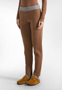 Bruine fleece broek met een grijze elastische tailleband. Heeft een taps toelopend ontwerp en zijzakken. Gedragen met bruin slippers en grijze sokken.