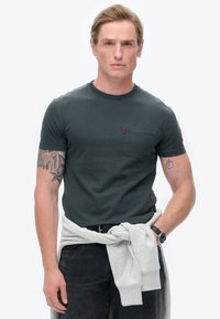 Mørkegrøn t-shirt med korte ærmer og en brystlomme, parret med grå træningsbukser der er bundet om livet. Modellen har lyse arm tatoveringer.