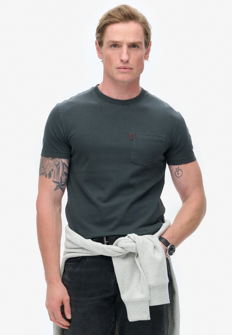 Mørkegrøn t-shirt med korte ærmer og en brystlomme, parret med grå træningsbukser der er bundet om livet. Modellen har lyse arm tatoveringer.