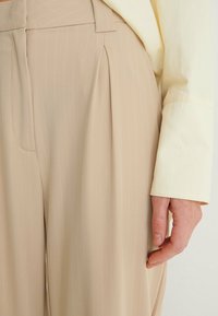 Beige veckade byxor med vertikala vita ränder, tillverkade av ett mjukt tyg. Byxorna har en sidoficka och en skräddarsydd passform.