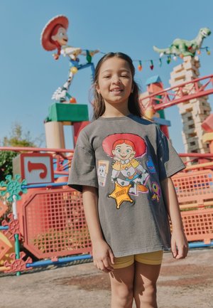 Ung pige iført en legetøjs sherifskjorte smiler udendørs ved en farverig Toy Story-temalegeplads med Woody og dinosaurfigurer.