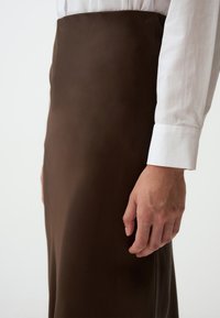 Gonna marrone realizzata in tessuto liscio, con design aderente e silhouette dritta, abbinata a una camicia bianca a maniche lunghe visibile nel fotogramma.