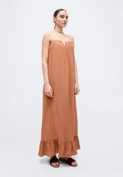 Kvinde med brunt hår bundet tilbage iført en stropløs maxi-kjole i rustfarve med flæsekant og sorte slip-on sandaler, stående mod en ensfarvet baggrund.