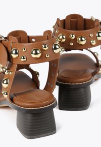 Ruske nahkalsandaler med gyldne metalstuds, en tykk svart hæl og justerbare ankelremmer med sirkulære utskjæringer.