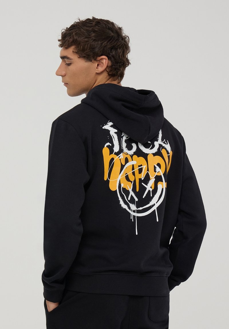 Młody mężczyzna w czarnej bluzie z kapturem z napisem "Za bardzo szczęśliwy" i graficznym uśmiechem z krzyżykami zamiast oczu z tyłu, stoi bokiem z rękami w kieszeniach.