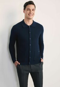 Granatowy sweter z dzianiny z kołnierzykiem, zapięciem na guziki i dwiema przednimi kieszeniami, noszony na szarych spodniach. Gładka tekstura i dopasowany krój.