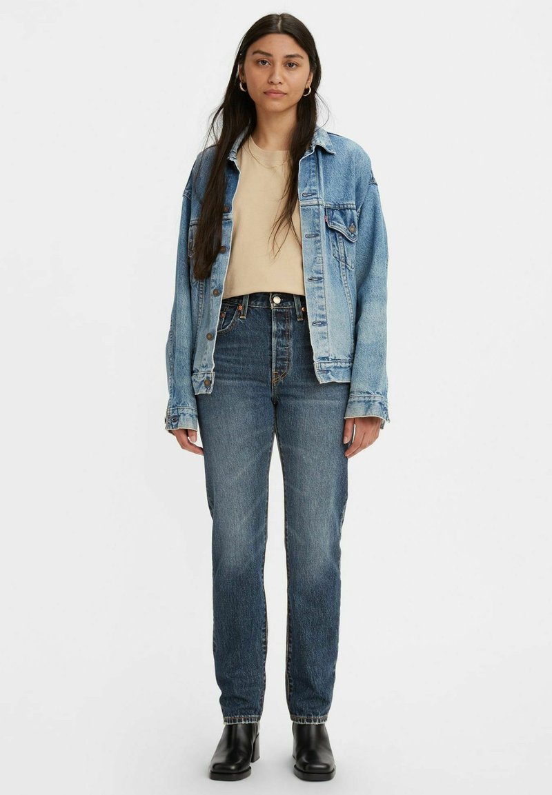 Mujer de pie, vistiendo una chaqueta de mezclilla azul claro sobre una camisa beige, jeans oscuros de tiro alto y botas negras de tobillo sobre un fondo blanco.