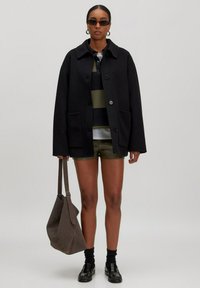 Schwarze oversized Wollmischung Jacke mit einem Kragen, vorderem Knopfverschluss und zwei Taschen. Kombiniert mit einem gestreiften Hemd und olivgrünen Shorts.