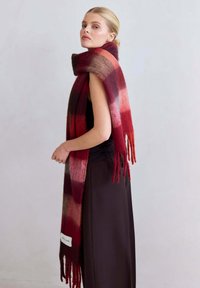 Écharpe en laine à carreaux rouges et bruns avec des franges aux extrémités, présentée drapée sur l'épaule. Étiquette visible en bas.