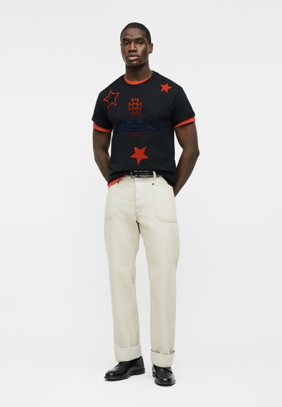 Άνδρας στέκεται με τα χέρια πίσω από την πλάτη, φοράει μαύρο μπλουζάκι με κόκκινα αστέρια και μπεζ παντελόνι με μαύρα παπούτσια, μπροστά σε λευκό φόντο.