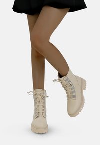 Bottes montantes à lacets de couleur crème avec une texture lisse et un accent argenté sur les côtés, dotées d'une semelle en caoutchouc robuste et d'un bout rond.