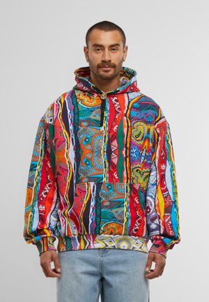 Man wearing a vibrant, multicolored patterned hoodie with a front pocket and light blue jeans against a plain background.