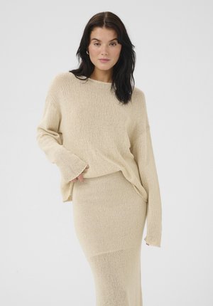 Beige gebreide trui en rok set. De trui heeft een losse pasvorm met wijde mouwen; de rok is aansluitend, beide zijn gemaakt van een textuurstof.