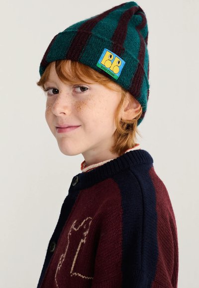 Enfant roux avec des taches de rousseur portant un bonnet rayé vert et marron et un pull sombre avec un motif d'ours, regardant de côté avec un léger sourire.
