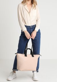 Duża jasnoróżowa torba tote z czarnymi uchwytami, gładka faktura, minimalistyczny design i przestronne wnętrze. Modelka trzyma torbę w codziennej stylizacji.