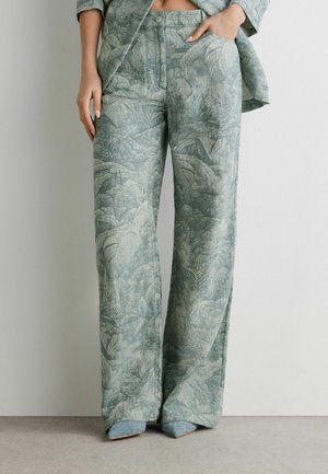 Donna che indossa pantaloni a gamba larga a vita alta con stampa botanica verde, abbinati a un blazer coordinato e scarpe intrecciate con punta a punta.