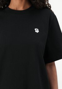 Camiseta de algodón negra con un pequeño bordado de flor blanca en la parte izquierda del pecho. Manga corta y escote redondo.