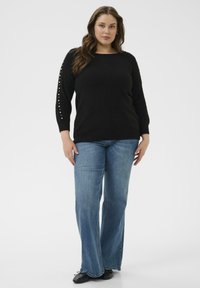 Schwarzer Strickpullover mit langen Ärmeln, der Perlendetails an den Ärmeln aufweist, kombiniert mit weit geschnittenen blauen Jeans und schwarzen Schuhen.