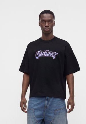 Jeune homme portant un t-shirt noir ample avec un texte violet stylisé et un jean en denim bleu clair sur un fond blanc uni.