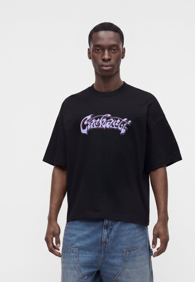 Junger Mann trägt ein lockeres schwarzes T-Shirt mit stilisiertem lila Text und hellblauen Jeans vor einem schlichten weißen Hintergrund.