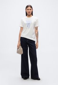 Λευκό βαμβακερό μπλουζάκι με μπλε γραφικό σχέδιο, συνδυασμένο με φαρδιά σκούρα τζιν παντελόνια. Άτομο που κρατάει μια μπεζ τσάντα με πτυχώσεις.