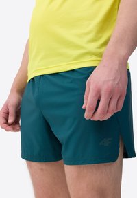 Teal sportshorts met een gladde textuur, met zijsplitten voor bewegingsvrijheid en een subtiele logo print. Gecombineerd met een felgeel mesh top.