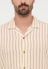 Άνδρας με κοντή γενειάδα που φοράει πουκάμισο με κάθετες ρίγες σε μπεζ και ανοιχτό καφέ, με κουμπιά και γιακά τύπου καμπ.