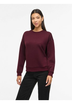 Naine, kes kannab pikkade varrukatega, ümara kaelusega burgundia tooni dressipluusi ja musti pükse, seisab ühevärvilise valge tausta ees.