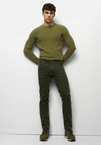 Pull en maille vert olive et jean côtelé vert olive foncé. Le mannequin se tient droit, portant des baskets sombres. Fond simple et minimal.