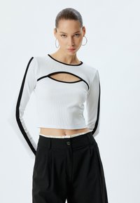 Witte lange-mouwen crop top met uitsnijdingen, zwarte accentrand langs de mouwen en halslijn, geribbelde textuur, gecombineerd met zwarte high-waisted broek.