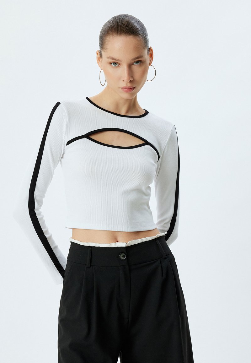 Witte lange-mouwen crop top met uitsnijdingen, zwarte accentrand langs de mouwen en halslijn, geribbelde textuur, gecombineerd met zwarte high-waisted broek.