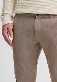 Hnědé strukturované kalhoty s jemným vzorem, vybavené předním zapínáním, bočními kapsami a kontrastním prošíváním.