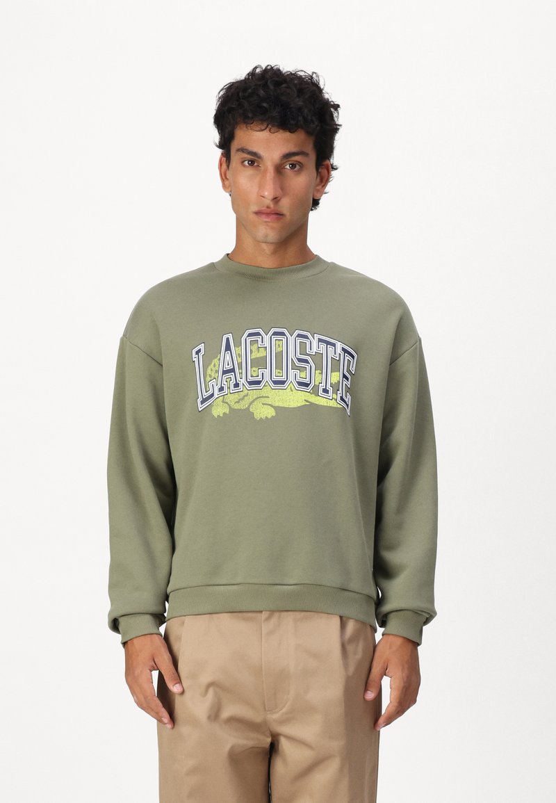 Sudadera verde oliva con cuello redondo acanalado, que presenta un gran logo de "LACOSTE" y un gráfico de cocodrilo en amarillo. Material de algodón.