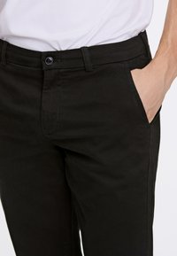 Pantaloni neri in cotone con chiusura a bottone, tasche laterali e una texture liscia. Cuciture visibili lungo le cuciture.