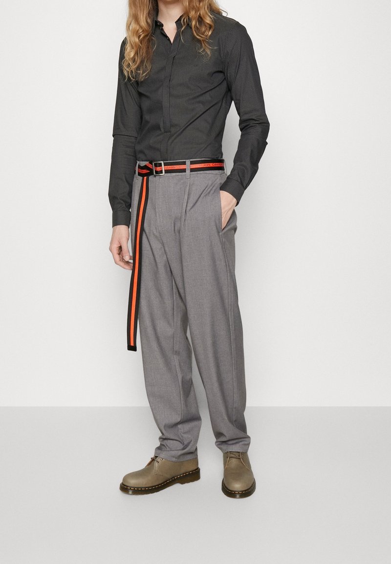 Grijze broek met een losse pasvorm en plooien, gecombineerd met een donkergeruit overhemd. De riem heeft een oranje streep op een zwarte achtergrond. Bruine schoenen.