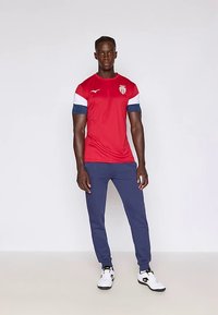 Červený sportovní dres s bílými a modrými akcenty a logem. Kombinován s tmavě modrými sportovními kalhotami a bílými teniskami.