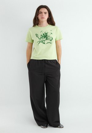 Kobieta stojąca z rękami w kieszeniach, ubrana w jasnozielony t-shirt z ciemnym kwiatowym nadrukiem i luźne czarne spodnie na prostym białym tle.