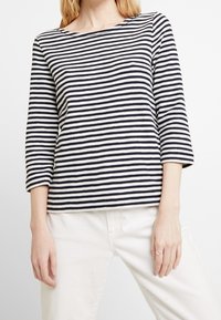 Top a righe navy e bianche, con maniche lunghe, realizzato in tessuto testurizzato, caratterizzato da un scollo ampio e maniche di mezza lunghezza. Indossato con pantaloni bianchi.