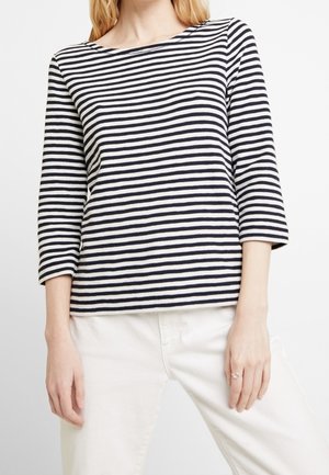 Top a righe navy e bianche, con maniche lunghe, realizzato in tessuto testurizzato, caratterizzato da un scollo ampio e maniche di mezza lunghezza. Indossato con pantaloni bianchi.