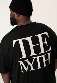 Černé oversize tričko s velkým bílým textem "THE MYTH" na zádech. Bavlněný materiál s měkkou texturou a volným střihem.