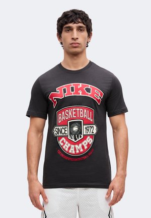 Νέος άνδρας που φοράει μαύρο t-shirt Nike με κόκκινο και λευκό λογότυπο πρωταθλητών μπάσκετ και λευκό αθλητικό σορτς σε απλό φόντο.