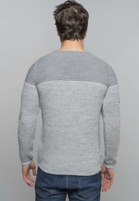 Pull en tricot gris avec un motif texturé, comportant une section supérieure côtelée et des manches ajustées. Porté avec un jean en denim bleu.