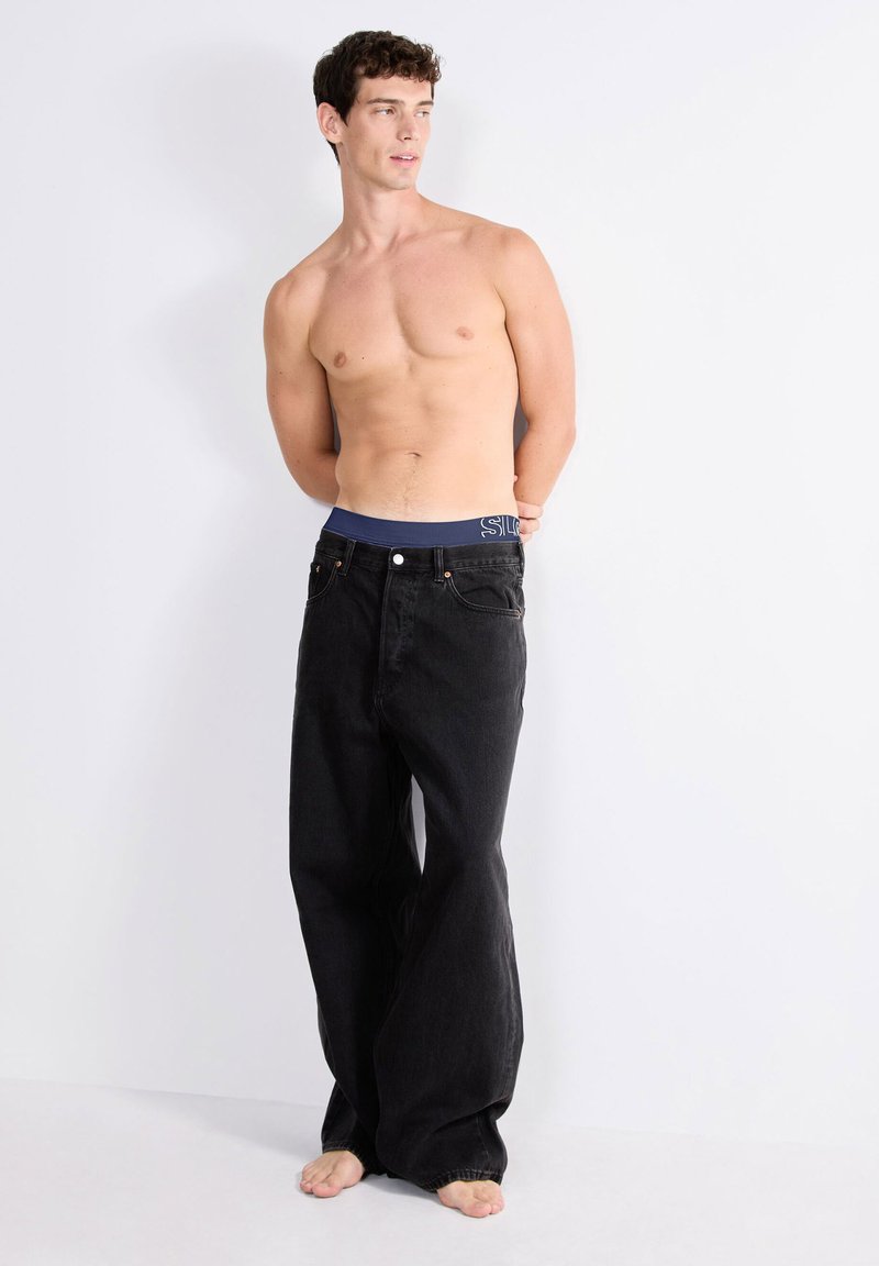 Shirtloze man met kort donker haar, gekleed in losse zwarte jeans en marineblauwe onderbroek, staande op blote voeten tegen een witte muur.