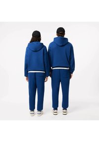 Blauwe hoodies met voorzakken en een trekkoordcapuchon, gecombineerd met bijpassende blauwe sweatpants. Beide outfits hebben een losse pasvorm.
